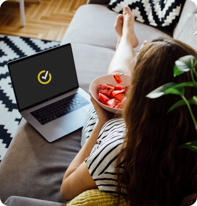 Kvinne som ligger på en sofa og spiser frukt fra en skål. Ved siden av henne er en bærbar PC som viser Norton-logoen