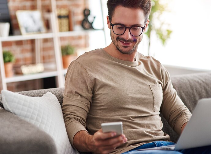 Mann som sitter på en sofa og ser på telefonen sin mens han har en bærbar PC i fanget